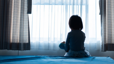 Young child sitting alone on bed looking out window, representing childhood injury victims and their families
