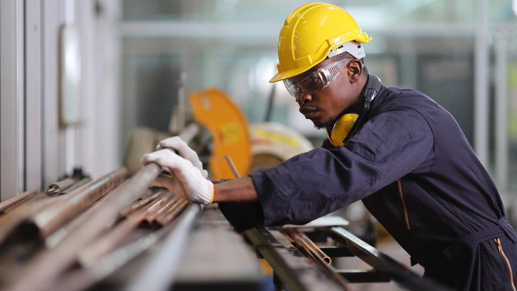 Factory worker in yellow hard hat and safety glasses handling copper pipes on industrial workbench