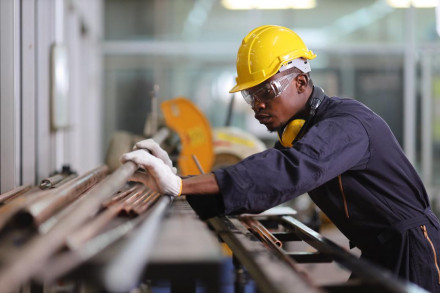 Factory worker in yellow hard hat and safety glasses handling copper pipes on industrial workbench