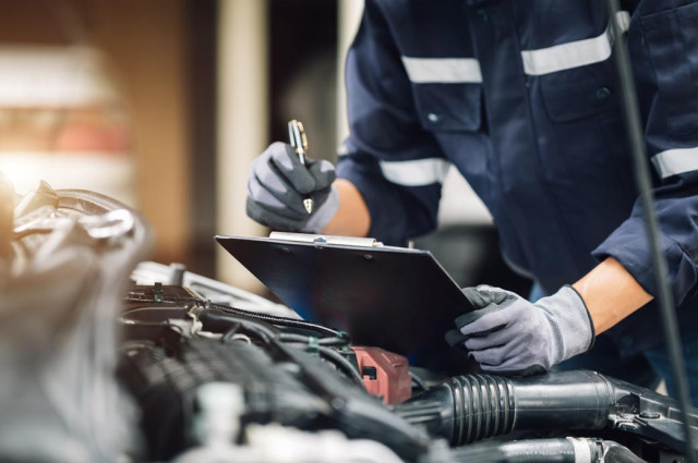 Mechanic in uniform inspecting vehicle engine components with clipboard at auto repair facility