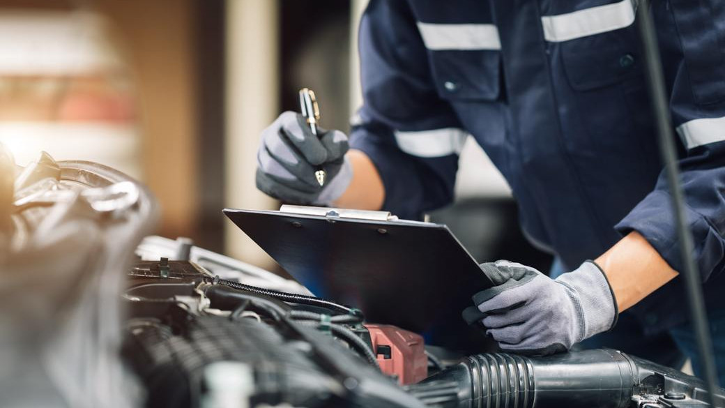 Mechanic in uniform inspecting vehicle engine components with clipboard at auto repair facility