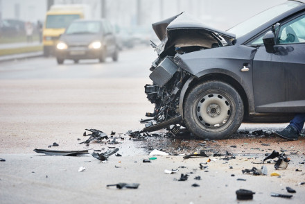 Crashed gray sedan with destroyed front end and debris scattered across a foggy urban roadway