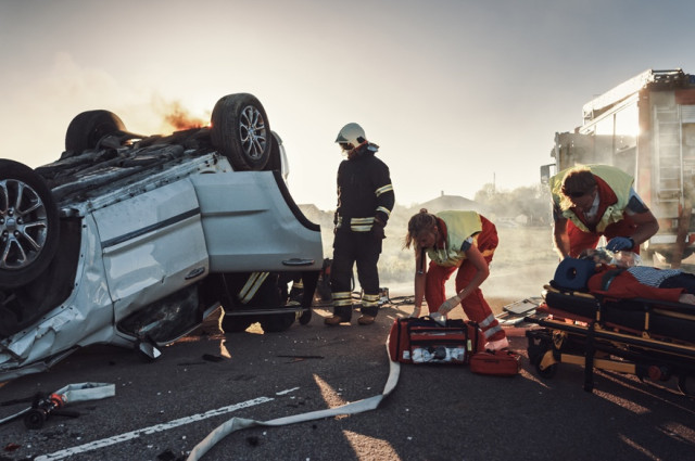 Paramedics treating an injured victim on a stretcher beside an overturned burning vehicle at a car accident scene