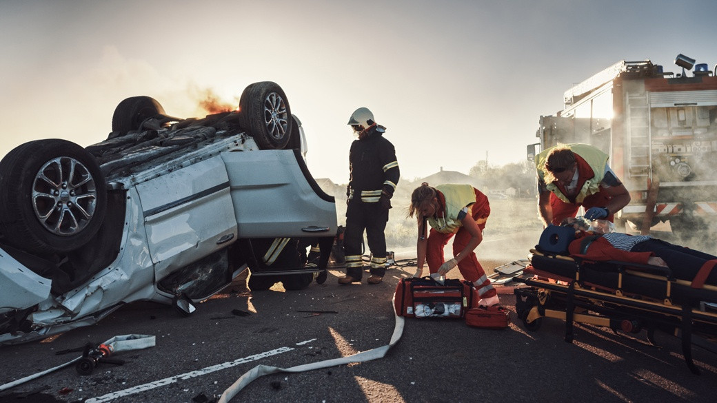 Paramedics treating an injured victim on a stretcher beside an overturned burning vehicle at a car accident scene