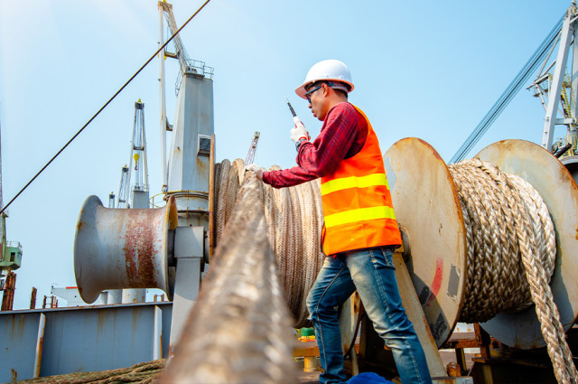Shipyard worker in hard hat and safety vest inspecting mooring lines on commercial vessel deck