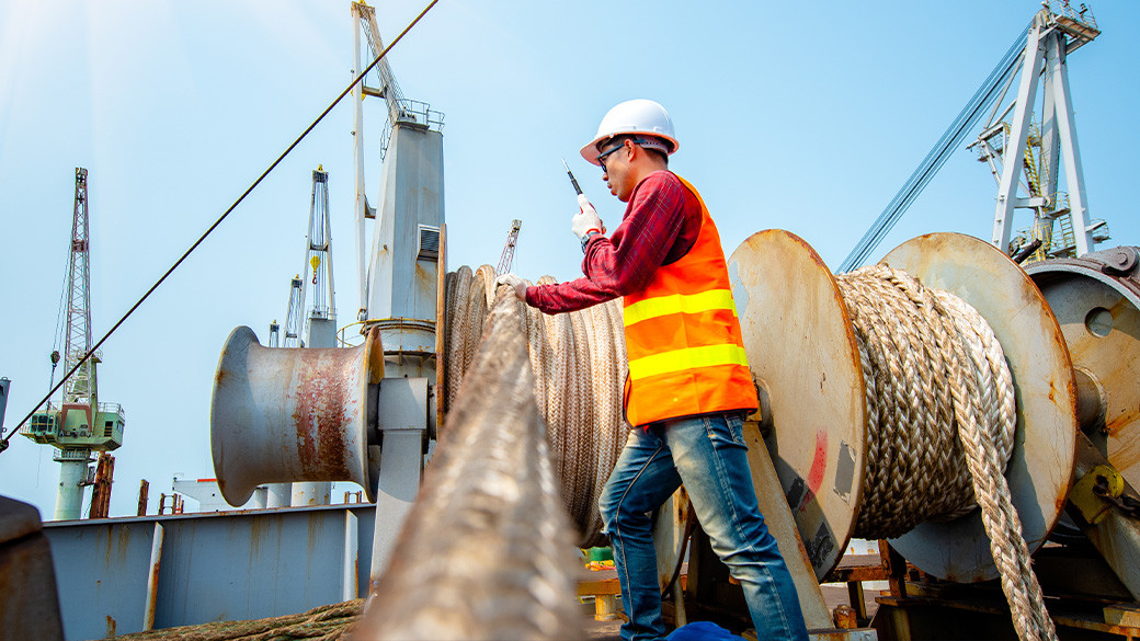 Shipyard worker in hard hat and safety vest inspecting mooring lines on commercial vessel deck