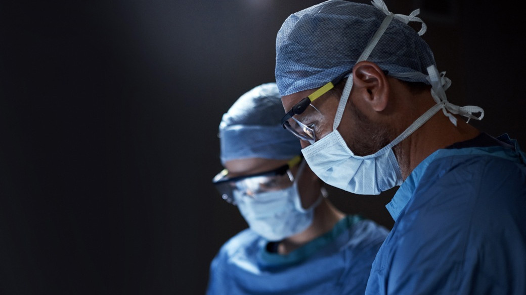 Two surgeons in profile wearing blue scrubs and protective eyewear concentrating during a medical procedure