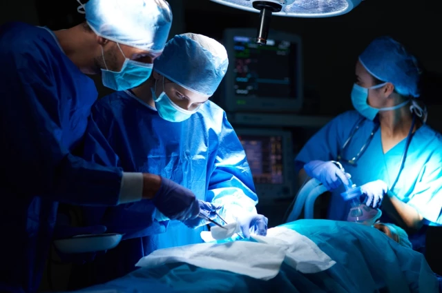 Three-person surgical team performing a procedure with anesthesiologist present in a hospital operating room