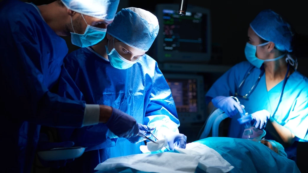 Three-person surgical team performing a procedure with anesthesiologist present in a hospital operating room